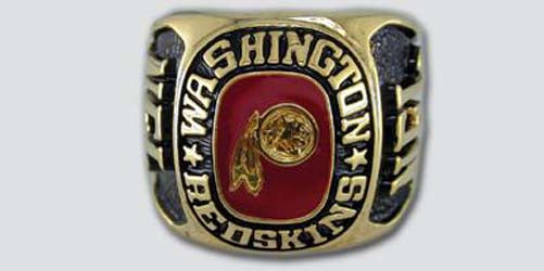 A gold colored paperweight featuring the Washington Redskins logo set against a red enamel background. The words "Washington Redskins" are engraved on the bezel.