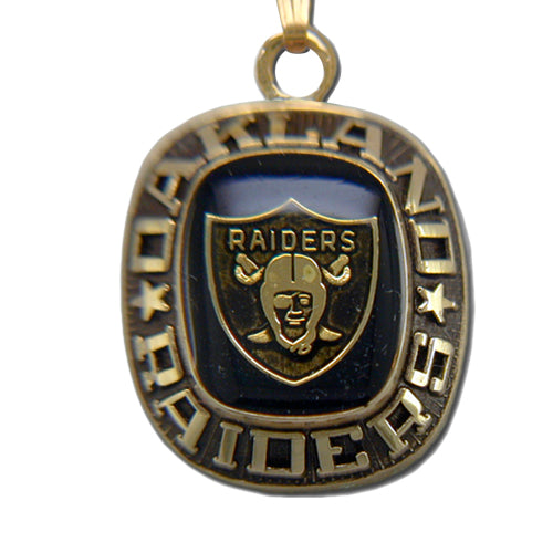 The Oakland Raiders Goldtone Pendant with Enamel features the team logo, “OAKLAND” above and “RAIDERS” below a black shield with helmet and swords—an eye-catching NFL accessory for any fan.