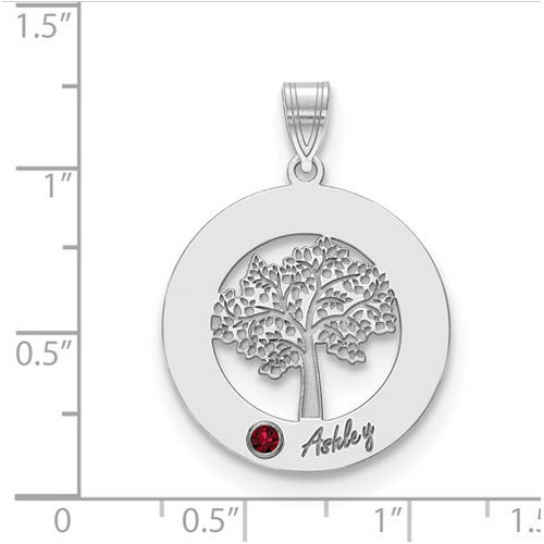 The Circle Pendant With 1 Birthstone showcases a round silver pendant featuring a detailed tree cutout. A small red birthstone is set at the bottom left of the tree, with the name "Ashley" inscribed nearby. This high-quality gold pendant hangs from a decorated bail, which has the letter "A" on its left side. The pendant is shown on a ruled background.