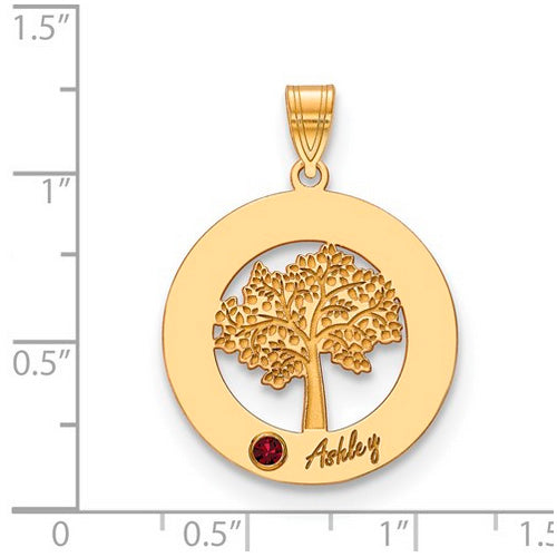 The Circle Pendant With 1 Birthstone showcases a round gold pendant featuring a detailed tree cutout. A small red birthstone is set at the bottom left of the tree, with the name "Ashley" inscribed nearby. This high-quality gold pendant hangs from a decorated bail, which has the letter "A" on its left side. The pendant is shown on a ruled background.