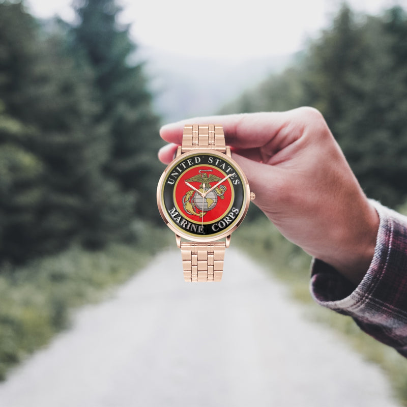 A hand in a plaid shirt holds the United States Marine Corps Goldtone Watch against the background of a blurred forest scene, where a path runs through the center.