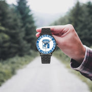 A hand in a plaid shirt holds the XVIII Airborne Corps Watch against the background of a blurred forest scene, where a path runs through the center.