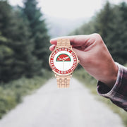 A hand in a plaid shirt holds the 78th Infantry Division Goldtone Watch against the background of a blurred forest scene, where a path runs through the center.