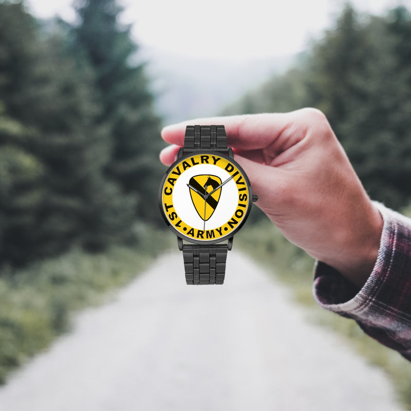 A hand in a plaid shirt holds the 1st Cavalry Division Watch against the background of a blurred forest scene, where a path runs through the center.