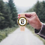 A hand in a plaid shirt holds the 101st Airborne Division Goldtone Watch against the background of a blurred forest scene, where a path runs through the center.