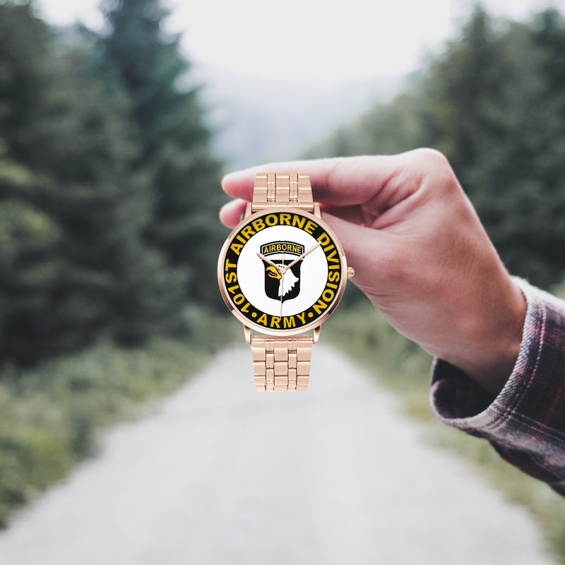 A hand in a plaid shirt holds the 101st Airborne Division Goldtone Watch against the background of a blurred forest scene, where a path runs through the center.