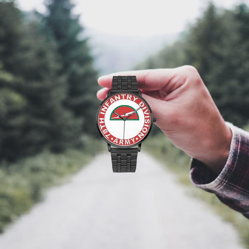 A hand in a plaid shirt holds the 78th Infantry Division Watch against the background of a blurred forest scene, where a path runs through the center.