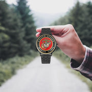 A hand in a plaid shirt holds the United States Marine Corps Watch against the background of a blurred forest scene, where a path runs through the center.