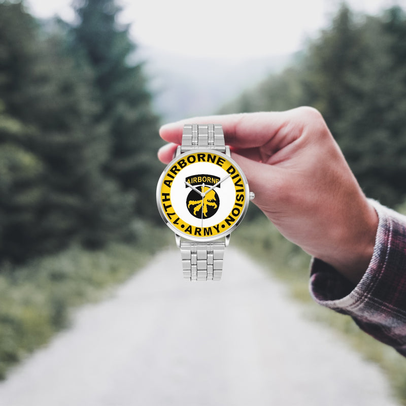 A hand in a plaid shirt holds the 17th Airborne Division Silvertone Watch against the background of a blurred forest scene, where a path runs through the center.