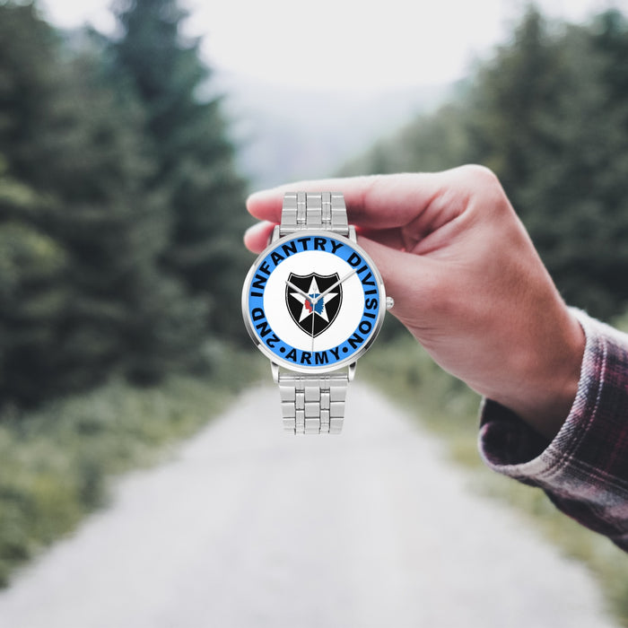 A hand in a plaid shirt holds the 2nd Infantry Division Silvertone Watch against the background of a blurred forest scene, where a path runs through the center.