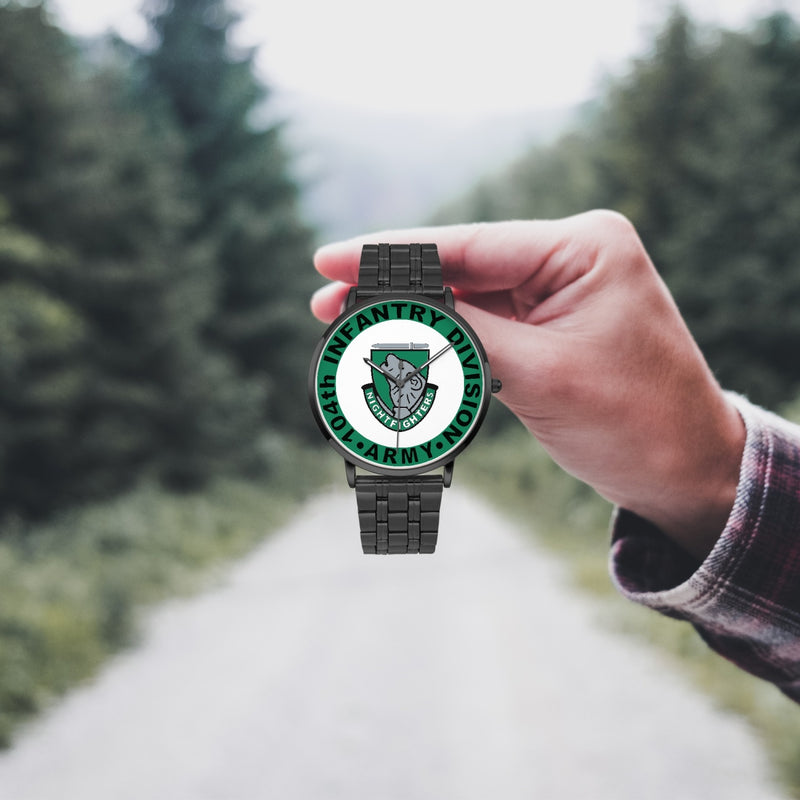 A hand in a plaid shirt holds the 104th Infantry Division Watch against the background of a blurred forest scene, where a path runs through the center.