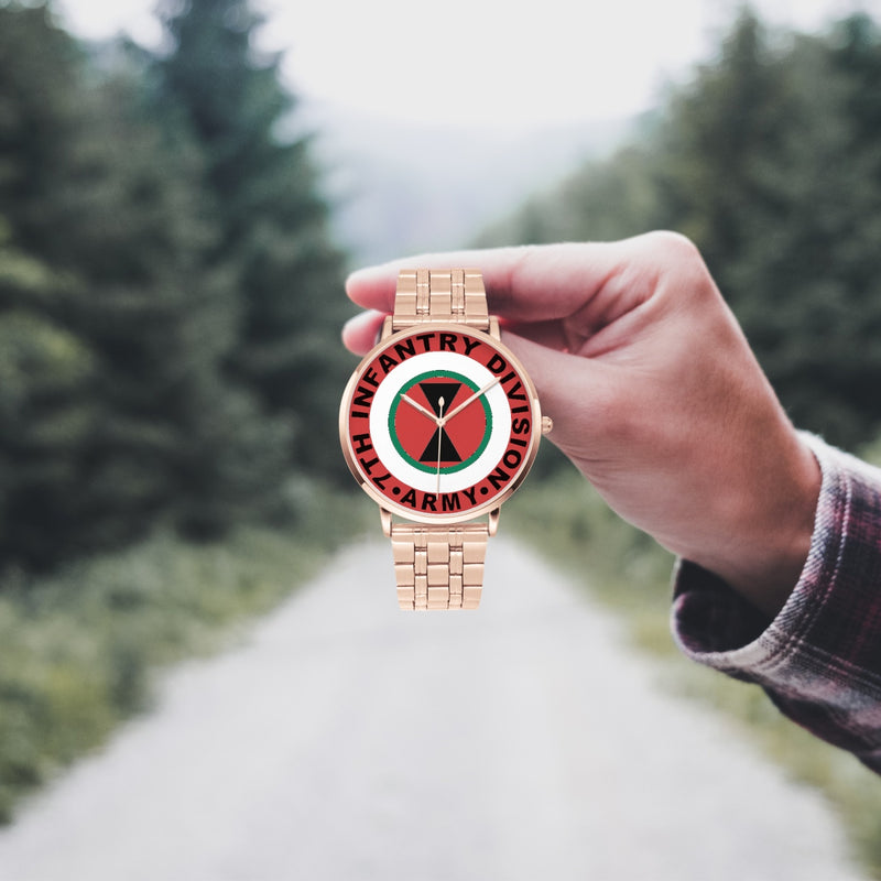 A hand in a plaid shirt holds the 7th Infantry Division Goldtone Watch against the background of a blurred forest scene, where a path runs through the center.