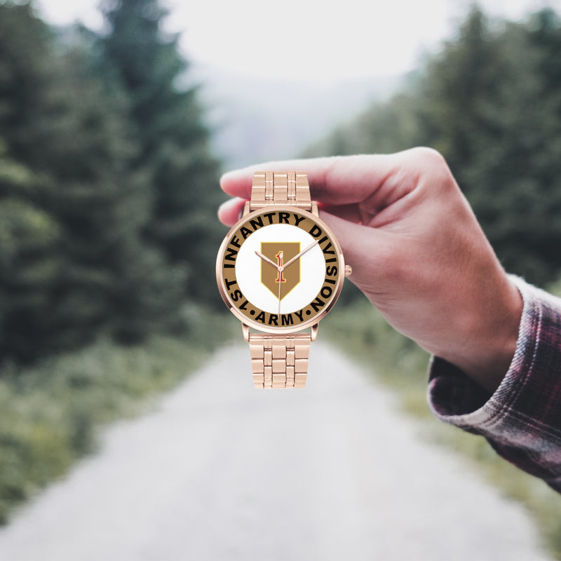 A hand in a plaid shirt holds the 1st Infantry Division Goldtone Watch against the background of a blurred forest scene, where a path runs through the center.