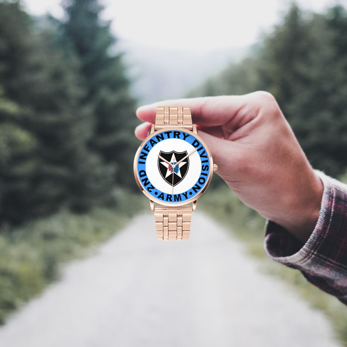 A hand in a plaid shirt holds the 2nd Infantry Division Goldtone Watch against the background of a blurred forest scene, where a path runs through the center.
