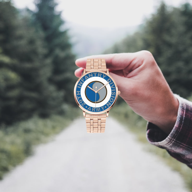 A hand in a plaid shirt holds the 29th Infantry Division Goldtone Watch against the background of a blurred forest scene, where a path runs through the center.