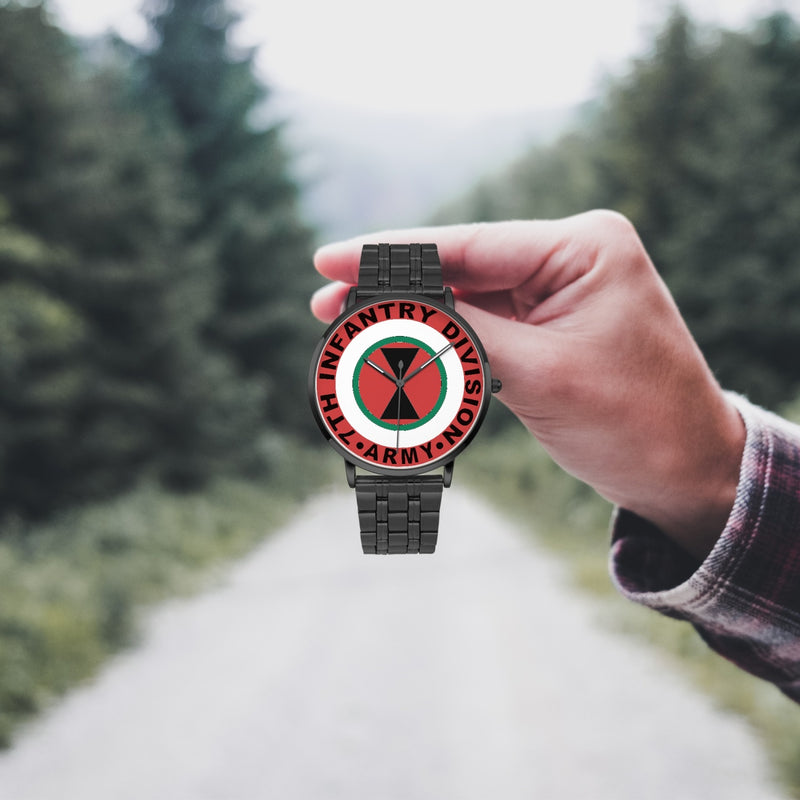 A hand in a plaid shirt holds the 7th Infantry Division Watch against the background of a blurred forest scene, where a path runs through the center.