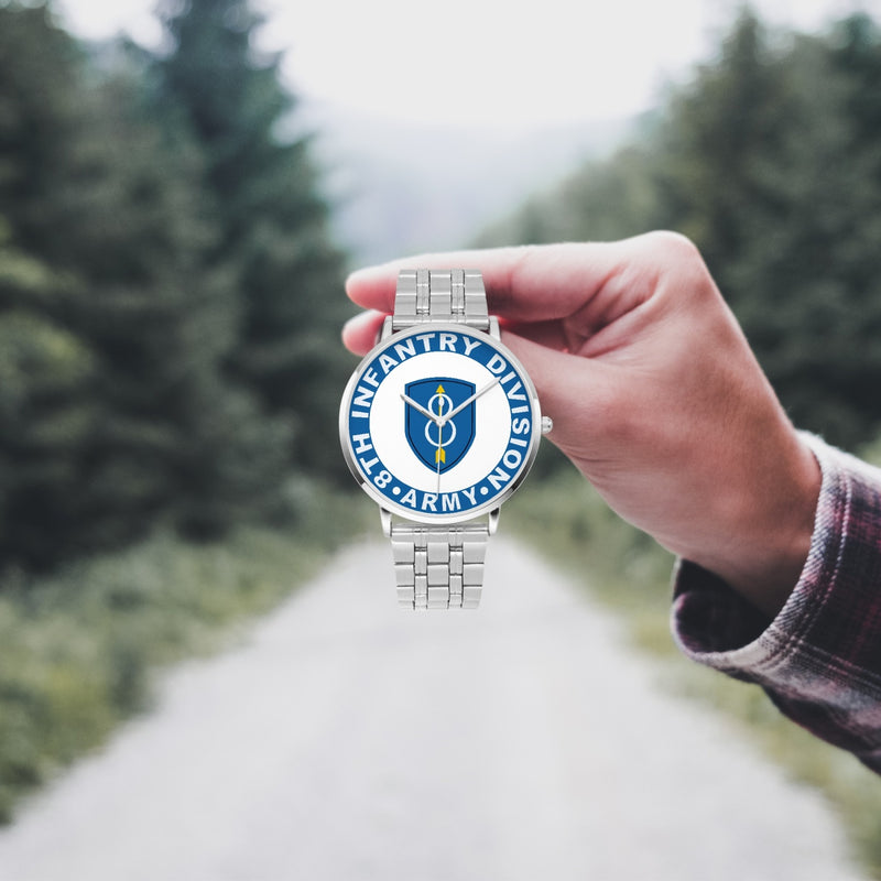 A hand in a plaid shirt holds the 8th Infantry Division Silvertone Watch against the background of a blurred forest scene, where a path runs through the center.