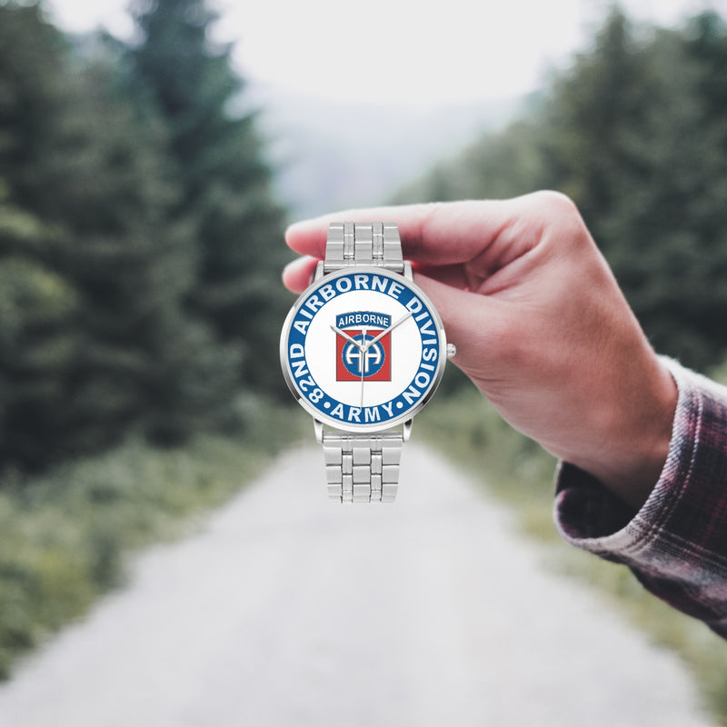A hand in a plaid shirt holds the 82nd Airborne Division Silvertone Watch against the background of a blurred forest scene, where a path runs through the center.