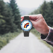 A hand in a plaid shirt holds the 42nd Infantry Division Watch against the background of a blurred forest scene, where a path runs through the center.
