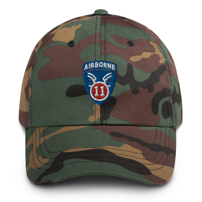 A military style 11th Airborne Division Hat with camouflage and the logo on the front.  The hat has a curved visor and an adjustable strap at the back. 